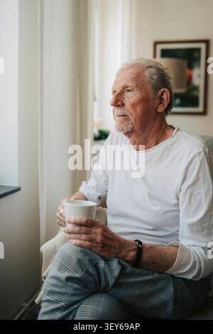 Thoughtful elderly man holding mug and looking away while sitting in retirement facility Stock Photo