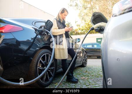 Woman charging her car at an ev station, using smartphone for sustainable transport Stock Photo