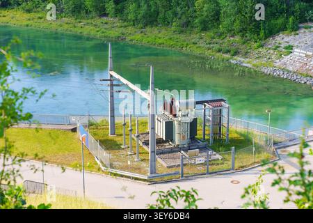 Electric power substation with a transformer stands fenced next to the turquoise waters of the Lech River in Schongau, Bavaria, Germany, surrounded by Stock Photo
