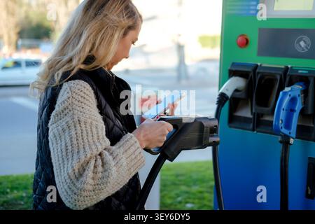 Woman standing beside charging station holding smartphone and electric car charger Stock Photo