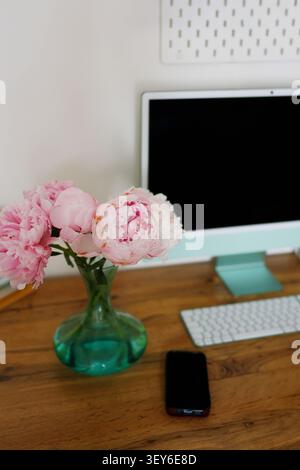 Modern home office workspace with blooming pink peonies, desktop computer and smartphone on a wooden desk in natural daylight. Stock Photo