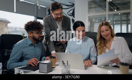 Multiracial business group four multiethnic professionals women men at office meeting briefing conference creating project with laptop papers smiling Stock Photo