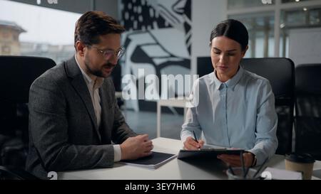 Two Caucasian business people office businesspeople woman and man talking analyzing notes documents papers businesswoman businessman colleagues Stock Photo