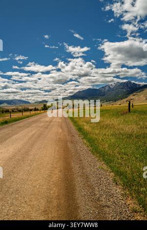 A wide angle vertical photo framed by blue sky, summer clouds, and distant hills curves through an open road through grassland. Stock Photo