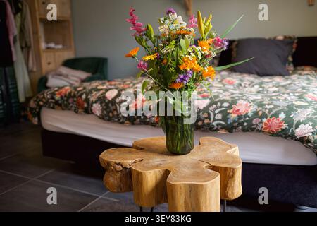 Bright mixed wildflower bouquet in clear glass vase on carved wood table with patterned bedspread  Stock Photo