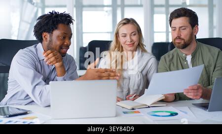 Three smiling professionals multiracial business colleagues talking about papers data information discussing trading startup report at boardroom Stock Photo