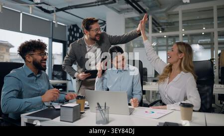 Four multiethnic business people working together with laptop talking winning job success giving high five celebrating multiracial businessmen and Stock Photo