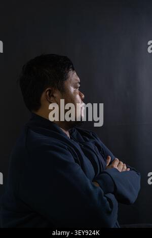 A thoughtful adult indonesian man stands in a side profile view with arms firmly crossed against a dark gray background, illuminated by moody lighting Stock Photo
