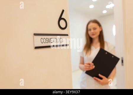 A close-up of a door sign reading COSMETOLOGY, featuring a blurred background of a female beauty technician or dermatologist holding a black clipboard Stock Photo
