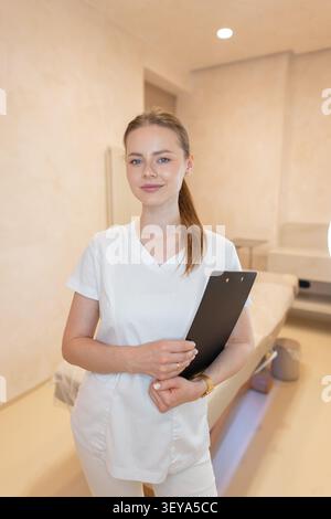 Friendly female cosmetologist, nurse or doctor in white medical uniform standing with black clipboard in a modern spa beauty clinic room. Smiling port Stock Photo