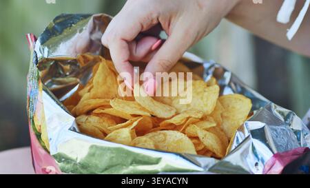 Woman's hand with pink nail polish picking a potato chip from an opened foil bag of chips Stock Photo