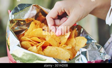 Woman munching crispy potato chip, pulling snack from opened bag while lounging, indulging salty treat with pleasure Stock Photo