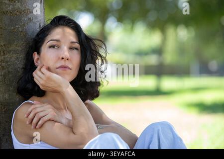 Young woman sitting under a tree in a peaceful park looking thoughtfully into the distance. Natural outdoor lifestyle portrait representing wellbeing, Stock Photo