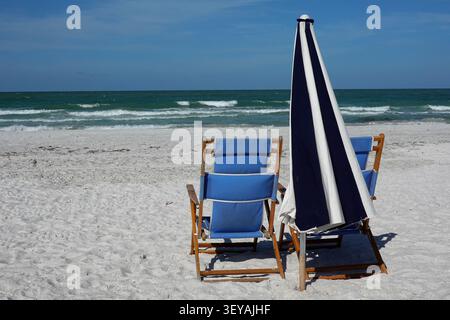 Blue beach chair with wooden frame next to a closed blue and white umbrella on a sandy beach. Stock Photo