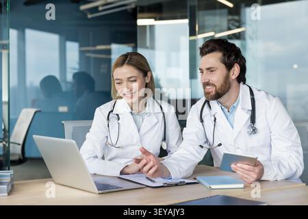 Two smiling doctors, a man and a woman, wearing lab coats and stethoscopes, collaborating on a laptop and holding a digital tablet while working in a modern clinic office Stock Photo