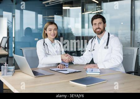 Two doctors. A man and a woman. Wearing white coats and stethoscopes. Shaking hands over a conference table in a modern office. Signifying professional agreement and successful teamwork in healthcare Stock Photo