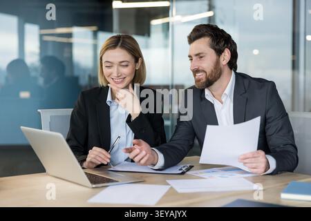 Two business colleagues are smiling and collaborating, reviewing financial data on a laptop and paper documents, discussing project strategy during a formal meeting in a contemporary workspace Stock Photo