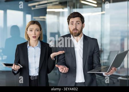 Two business coworkers standing in a modern office, one holding a tablet and the other a laptop, expressing confusion and doubt with open hands and shrugging shoulders Stock Photo