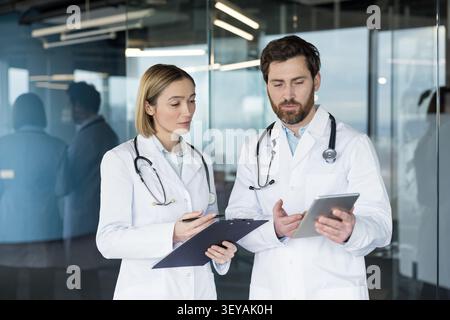 Two medical professionals collaborating and reviewing patient information on a digital tablet and physical clipboard in a modern office environment Stock Photo