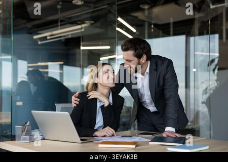 Diverse business professionals working together in a contemporary office setting, a man supporting a woman sitting at her desk, discussing work with positive expressions Stock Photo