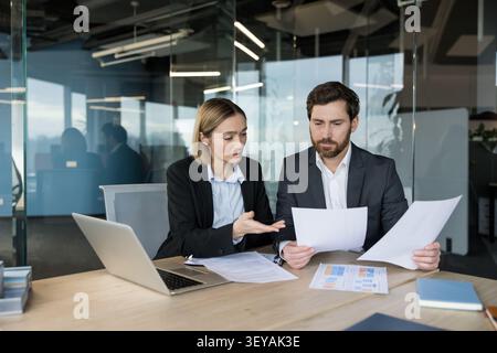 Two business colleagues are sitting at a desk in a modern office, discussing documents and analyzing data while using a laptop for project planning and professional collaboration Stock Photo