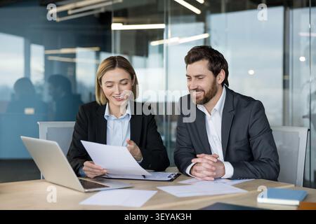 Two smiling business professionals working together at a desk, reviewing documents and a laptop, discussing corporate strategy and partnership in a contemporary office setting Stock Photo