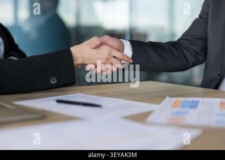 Business partners finalizing a deal by shaking hands over a meeting desk with documents and a pen, symbolizing agreement, partnership, success, and trust in a professional office setting Stock Photo