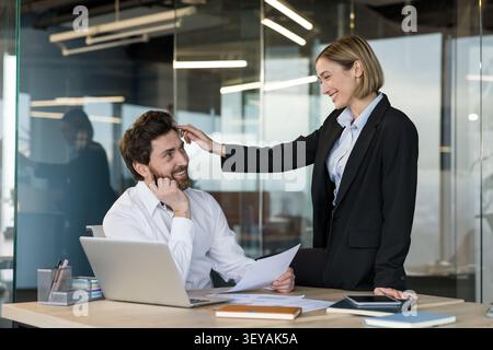 Happy business colleagues engaging in playful professional interaction, a woman gently touching a man's hair at his desk, creating a concept of office romance or flirtation Stock Photo