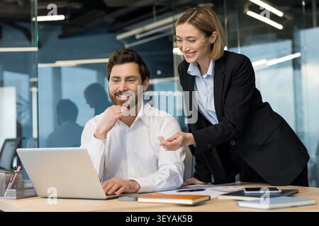 Business colleagues smiling while collaborating on a project, discussing new strategy and solutions using a laptop at their desk in a contemporary corporate office environment Stock Photo
