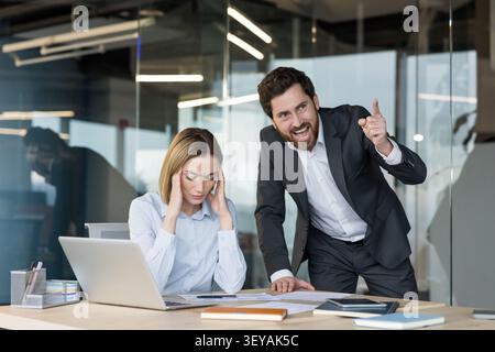 Angry businessman shouting and scolding his female employee, creating a toxic workplace environment, causing stress and emotional distress to the woman sitting at her desk Stock Photo