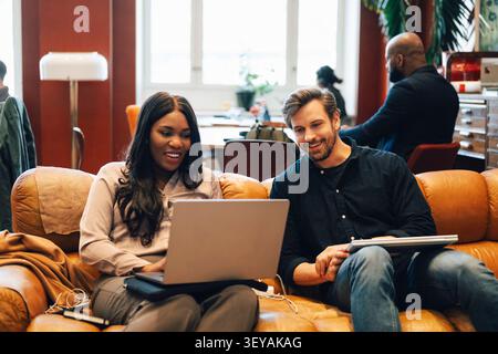 Smiling male and female business professionals discussing project on laptop while sitting on sofa in coworking space lou Stock Photo