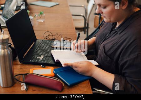 Female freelancer taking down notes in diary while sitting with laptop at shared working desk Stock Photo