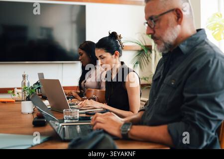 Group of freelancers working independently on laptops while sitting side by side at coworking space desk Stock Photo