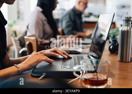 Close-up female freelancer working on laptop next to cup of tea at shared working desk Stock Photo