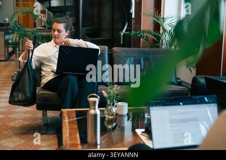 Female entrepreneur holding her bag while sitting with open laptop on sofa in coworking space lounge Stock Photo
