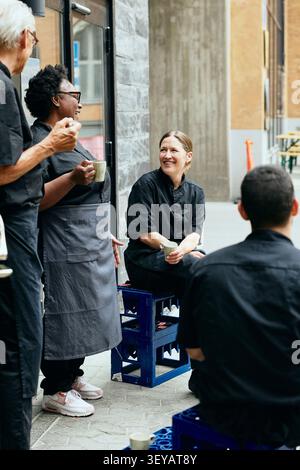 Diverse restaurant staff in black uniforms talking and drinking coffee while sitting outside back door Stock Photo