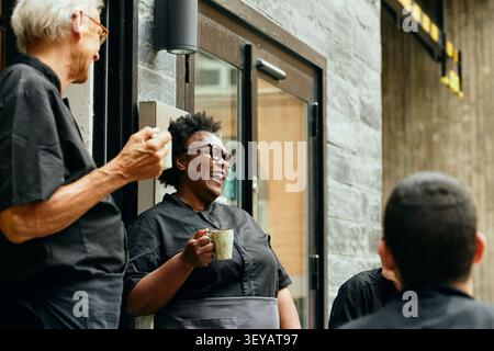 Cheerful female chef laughing with colleagues during outdoor coffee break Stock Photo