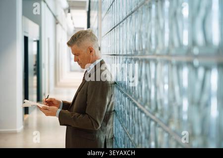 Side view of senior businessman analyzing reports while leaning on glass brick wall Stock Photo