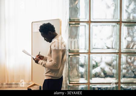 Side view of businessman writing on documents while leaning on glass brick wall Stock Photo