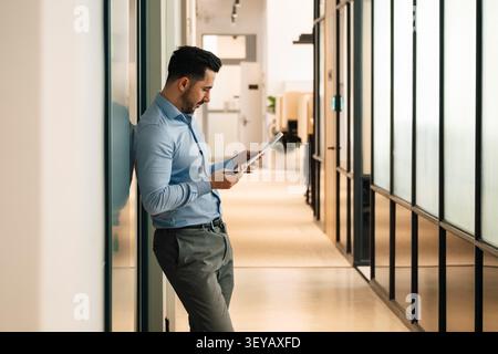 Businessman analyzing reports while leaning on wall in office hallway Stock Photo