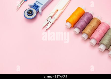 Sewing essentials including colorful thread spools, thread snips, rotary cutter, and seam ripper arranged on a pastel pink background with copy space, Stock Photo