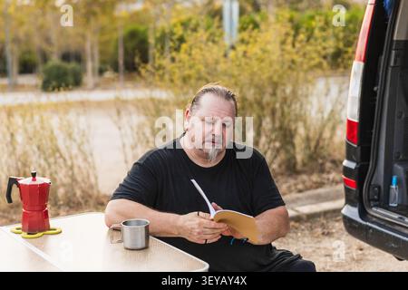 Mature man reading book while camping with van Stock Photo