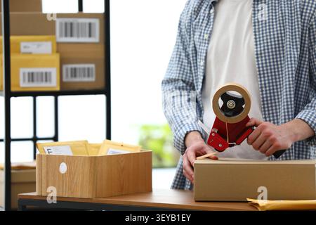 Man using dispenser with adhesive tape to seal cardboard box at table in office, closeup. Sending envelopes and parcels Stock Photo