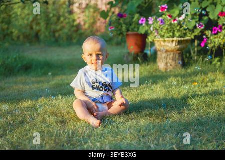 boy one year old boy sitting on the grass in the summer dressed in a T-shirt and diaper, baby European appearance blond with blue eyes. High quality Stock Photo