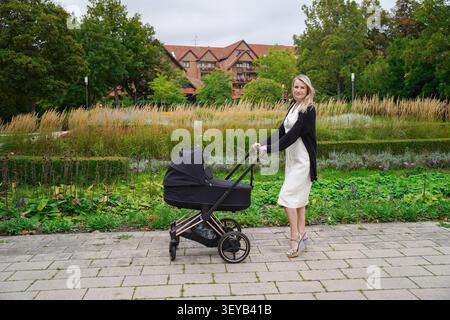 business mom dressed in a white dress on her lunch break pushing a black baby stroller on a bright fall day in the park, mom is a blonde European Stock Photo