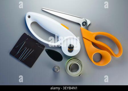 Sewing tools are arranged on a table including scissors, a rotary cutter, needles, a thimble, and a measuring tape. Stock Photo