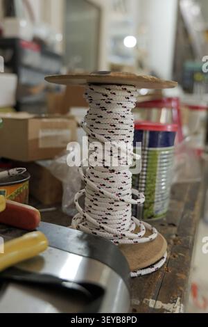 Rope spool with white and red speckled rope winding around a wooden core, sitting on a worn workbench in a workshop environment with various items blu Stock Photo