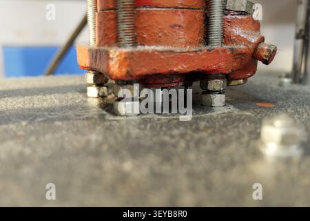 Close up detail of an old industrial machine's rusted red metal base firmly secured with nuts and bolts to a textured gray surface in a factory settin Stock Photo