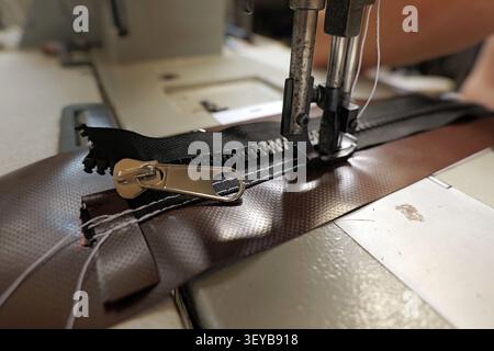 Industrial sewing machine stitching a black zipper onto heavy brown synthetic fabric, demonstrating the process of textile manufacturing and craftsman Stock Photo
