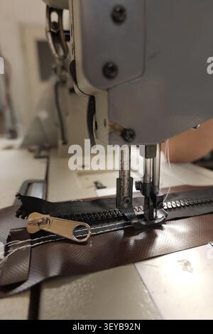 Industrial sewing machine mechanism working on a black zipper with a metallic slider. Attaching it to a brown textured synthetic fabric with white thr Stock Photo